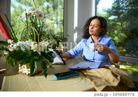 Charming woman floral designer holding a cup of coffee, sketching on digital tablet, working on new flower arrangements Charming woman floral designer holding a cup of coffee, sketching on digital tablet, working on new flower arrangements 104880778