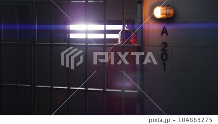 Prisoner sits on bed and looks on barred window 104883275