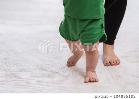 Cute little boy learning to make first steps with little legs on soft white rug in nursery caring mother holding hands of barefoot son teaching to walk at home closeup Cute little boy learning to make first steps with little legs on soft white rug in nursery caring mother holding hands of barefoot son teaching to walk at home closeup 104886185