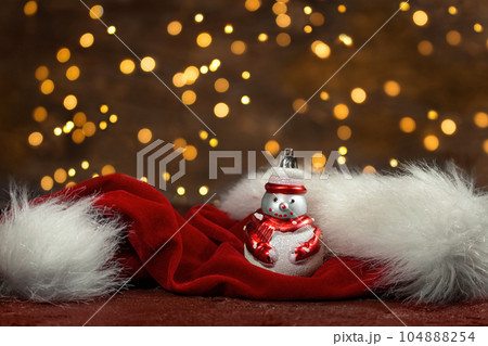 Snowman and red santa claus hat with new year lights in the back. Blurred background. Christmas card Snowman and red santa claus hat with new year lights in the back. Blurred background. Christmas card 104888254