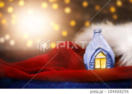 Decorative ice house on Santa Claus's red velvet hat with fur. Blurred lights with lens flare. Christmas, New Year postcard. Copy spac Decorative ice house on Santa Claus's red velvet hat with fur. Blurred lights with lens flare. Christmas, New Year postcard. Copy spac 104888256