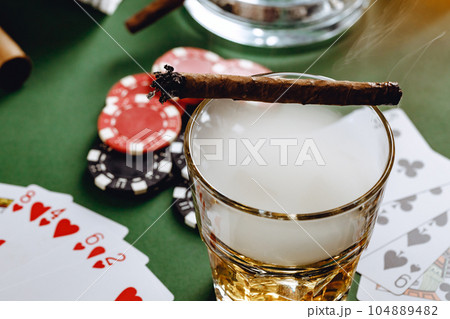 Glass of whiskey, cigar, playing cards and chips on green background Glass of whiskey, cigar, playing cards and chips on green background 104889482