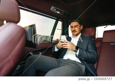 Young elegant businessman sitting on the back seat of a luxury car and using smartphone 104889727