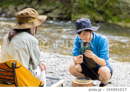 川原のキャンプで蚊取り線香をつける男性 104891017