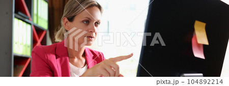 Business woman with serious face at office desk points her finger at computer screen. Business woman with serious face at office desk points her finger at computer screen. 104892214