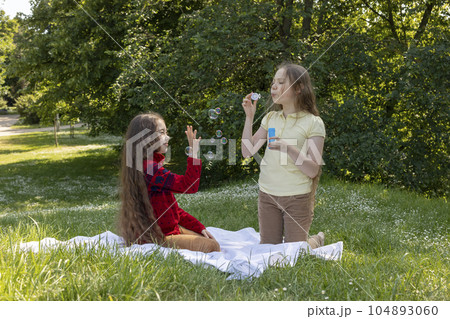 Joyful Asian Caucasian Girls, Children Of Elementary School Age Blow Soap Bubbles In Summer Park, Meadow. Happy Childhood. School Break Time. Children Protection Day, Earth Day. Horizontal Plane Joyful Asian Caucasian Girls, Children Of Elementary School Age Blow Soap Bubbles In Summer Park, Meadow. Happy Childhood. School Break Time. Children Protection Day, Earth Day. Horizontal Plane 104893060