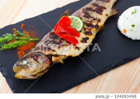 Tasty baked whole trout with rice, served with lemon and greens at plate Tasty baked whole trout with rice, served with lemon and greens at plate 104893498