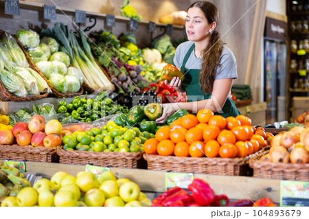 Female seller holding green peppers standing in fruit and vegetable section of supermarket Female seller holding green peppers standing in fruit and vegetable section of supermarket 104893679