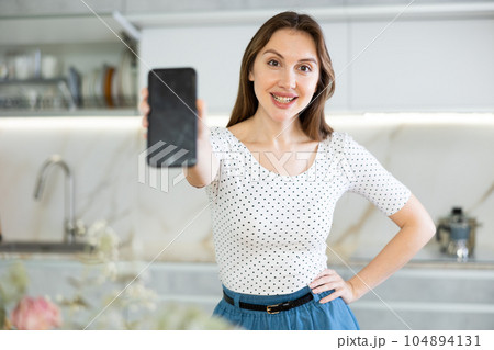 Woman student showing modern smartphone with blank screen in kitchen 104894131