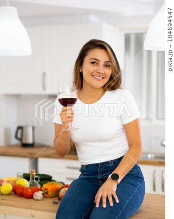 Smiling young Hispanic woman drinking wine sitting on table in kitchen Smiling young Hispanic woman drinking wine sitting on table in kitchen 104894547