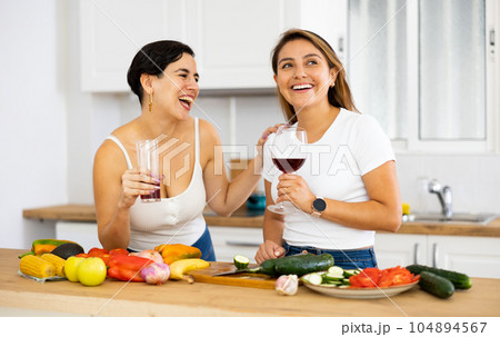 Happy female friends with glasses of red wine chatting and preparing salad together in kitchen Happy female friends with glasses of red wine chatting and preparing salad together in kitchen 104894567