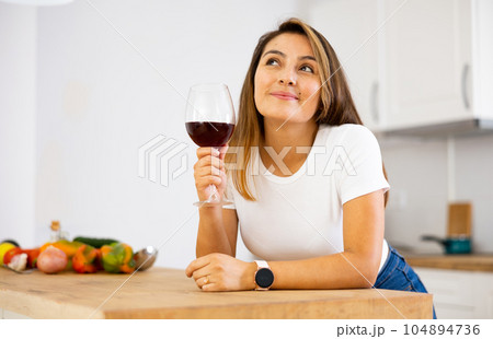 Portrait of woman with glass of red wine in modern kitchen - housewife prepares a vegetable salad and drinks red wine Portrait of woman with glass of red wine in modern kitchen - housewife prepares a vegetable salad and drinks red wine 104894736