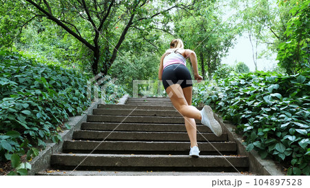 Beautiful sexy athletic young blond woman runs upstairs, in the forest, park, in summer day. Performs exercises to strengthen the different muscles of the legs on the stairs. Beautiful sexy athletic young blond woman runs upstairs, in the forest, park, in summer day. Performs exercises to strengthen the different muscles of the legs on the stairs. 104897528