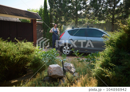 Young man in work overalls, using a water high pressure cleaners, washes his car in the backyard Young man in work overalls, using a water high pressure cleaners, washes his car in the backyard 104898942