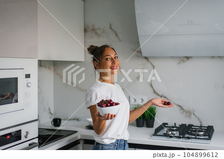 Pretty smiling young woman holds a plate of fresh cherry berries in her hand in the kitchen Pretty smiling young woman holds a plate of fresh cherry berries in her hand in the kitchen 104899612