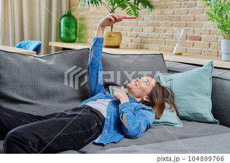 Young woman having video call lying on sofa at home, using smartphone Young woman having video call lying on sofa at home, using smartphone 104899766
