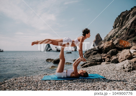 Woman sea yoga. Two Happy women meditating in yoga pose on the beach, ocean and rock mountains. Motivation and inspirational fit and exercising. Healthy lifestyle outdoors in nature, fitness concept. Woman sea yoga. Two Happy women meditating in yoga pose on the beach, ocean and rock mountains. Motivation and inspirational fit and exercising. Healthy lifestyle outdoors in nature, fitness concept. 104900776