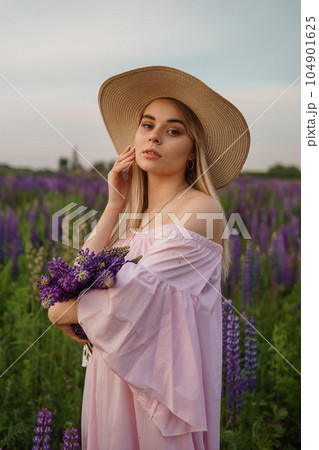A beautiful woman in a straw hat walks in a field with purple flowers. A walk in nature in the lupin field 104901625