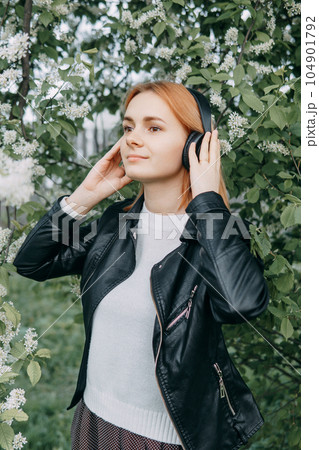 A red-haired teenage girl listens to music in headphones, configured via the phone. The time of spring flowering of cherry orchards A red-haired teenage girl listens to music in headphones, configured via the phone. The time of spring flowering of cherry orchards 104901792