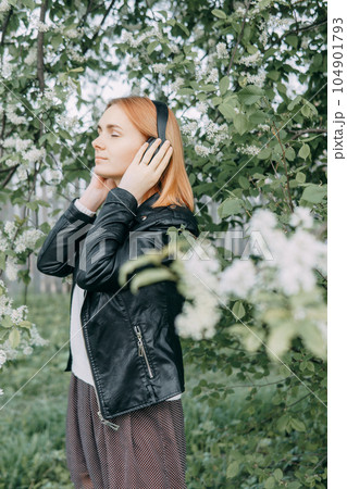 A red-haired teenage girl listens to music in headphones, configured via the phone. The time of spring flowering of cherry orchards A red-haired teenage girl listens to music in headphones, configured via the phone. The time of spring flowering of cherry orchards 104901793