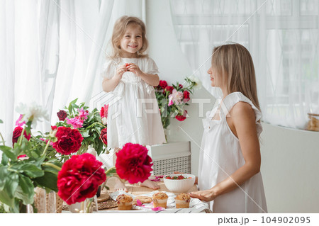A little blonde girl with her mom on a kitchen countertop decorated with peonies. The concept of the relationship between mother and daughter. Spring atmosphere. 104902095