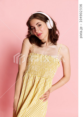Portrait of young beautiful girl in cute dress listening to music in headphones and smiling against pink studio background 104902336