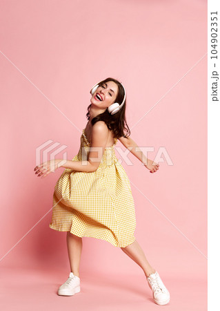 Happy young woman in yellow dress listening to music in headphones and dancing against pink studio background. Joy 104902351