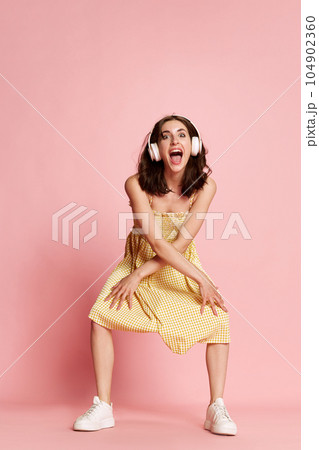 Smiling, young, beautiful girl in yellow dress listening to music in headphones and funny dancing against pink studio background 104902360