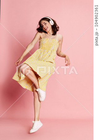 Happy, positive young pretty girl in yellow dress listening to music in headphones and dancing against pink studio background Happy, positive young pretty girl in yellow dress listening to music in headphones and dancing against pink studio background 104902361