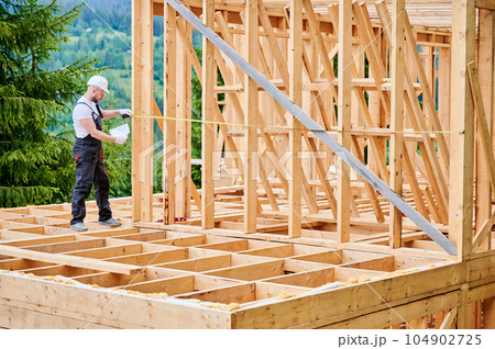 Carpenter building wooden skeleton house. Man measures distances with tape measure while wearing work attire and safety helmet. The principles of contemporary, eco-friendly building practices. 104902725