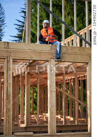 Carpenter constructing wooden frame two-story house near the forest. Bearded man holding hammer, dressed in protective helmet and orange safety vest. Concept of ecological modern construction. Carpenter constructing wooden frame two-story house near the forest. Bearded man holding hammer, dressed in protective helmet and orange safety vest. Concept of ecological modern construction. 104902730