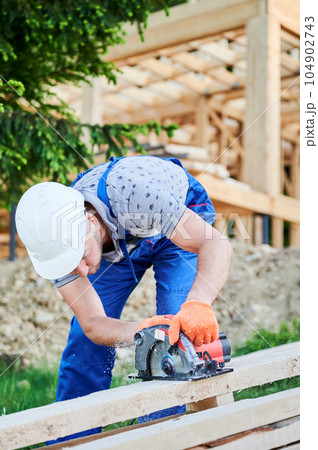 Carpenter using circular saw for cutting wooden plank. Man worker building wooden frame house. The ideology behind modern, environmentally-conscious construction. Carpenter using circular saw for cutting wooden plank. Man worker building wooden frame house. The ideology behind modern, environmentally-conscious construction. 104902743