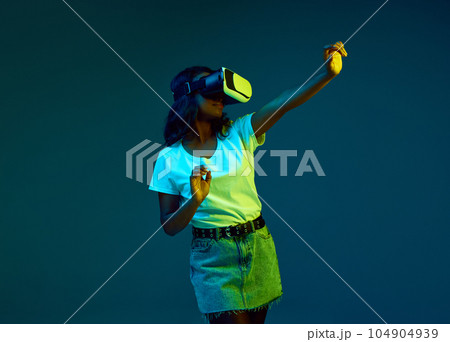 Portrait of attractive young, african woman in white t-shirt wearing vr glasses, posing against cyan, blue studio background in neon light 104904939