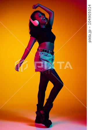 Full-length portrait of beautiful, young, african, stylish woman listening to music in headphones and dancing against yellow studio background in neon light 104905014