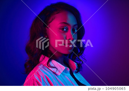 Close-up portrait of attrcative african woman looking at camera, posing against gradient blue purple studio background in neon light 104905065