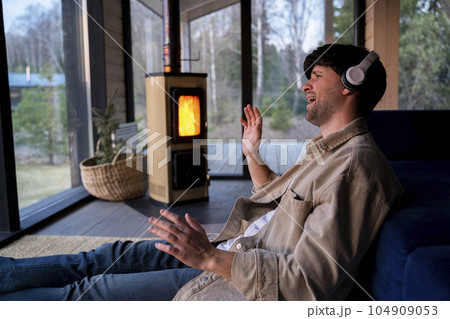 Energetic, positive guy sings loudly while listening to music with headphones, dressed in a beige shirt, sitting by the sofa in the living room against the background of the fireplace 104909053