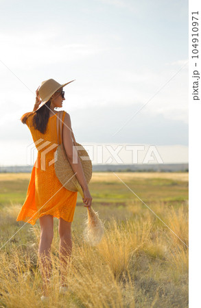 Beauty romantic girl outdoors. Rear view of a beautiful girl dressed in a casual orange dress with a straw hat and a straw bag in her hands in a field in the sunlight. Blows long hair. Autumn. Shine 104910971