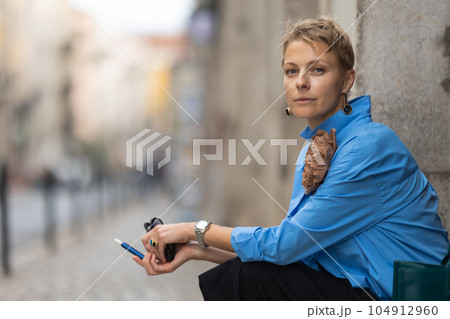 Adult stylish woman with short hair sitting on the european street and holding an electronic cigarette - looking in the camera 104912960