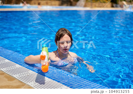 Young pretty caucasian girl with blue eyes shoots with a water gun in the pool. Pretty girl playing with water guns in pool. Cheerful children playing waterguns, rejoicing, jumping, swimming in pool. Young pretty caucasian girl with blue eyes shoots with a water gun in the pool. Pretty girl playing with water guns in pool. Cheerful children playing waterguns, rejoicing, jumping, swimming in pool. 104913255