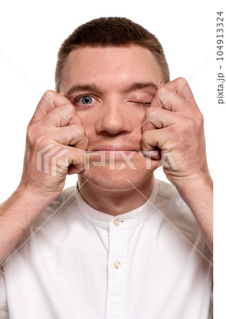 Charming handsome young man in a white shirt is making faces, while standing isolated on a white background Charming handsome young man in a white shirt is making faces, while standing isolated on a white background 104913324