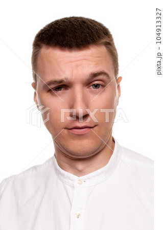 Charming handsome young man in a white shirt is making faces, while standing isolated on a white background Charming handsome young man in a white shirt is making faces, while standing isolated on a white background 104913327