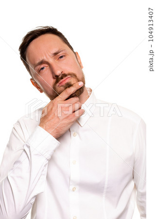 Portrait of unhappy determined european male with bristle touching chin while thinking and looking with serious and worried look at camera, standing against white background. 104913407