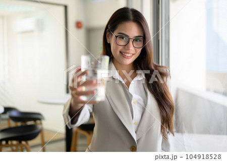 Confident businesswoman holding glass of water smiling to camera at office Confident businesswoman holding glass of water smiling to camera at office 104918528