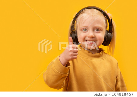Little blonde girl wears black headphones smiling and shows thumbs up. Portrait of preschool girl on yellow background. Copy space. 104919457