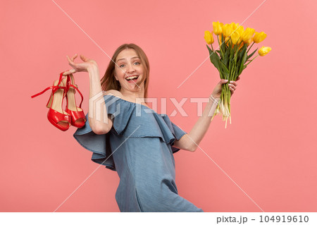 Happy fun young woman in summer sundress stands barefoot holds sandals and bouquet of flowers. Isolated on pink background. Copy space. Happy fun young woman in summer sundress stands barefoot holds sandals and bouquet of flowers. Isolated on pink background. Copy space. 104919610