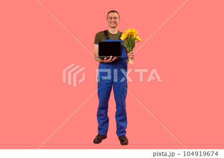 Full-length portrait of man in workers uniform holding laptop and bouquet of flowers, pink background. Copy space, mock up 104919674