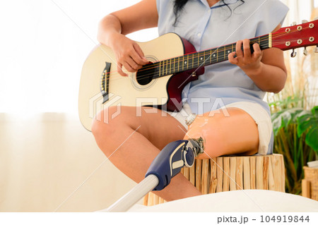 Young woman playing guitar and relaxing at home, Positive woman with prosthetic leg having fun on weekend at home, Leisure lifestyles and activities at home, Woman working at home office Young woman playing guitar and relaxing at home, Positive woman with prosthetic leg having fun on weekend at home, Leisure lifestyles and activities at home, Woman working at home office 104919844