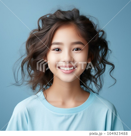 Portrait of a smiling Asian little girl with black hair on a blue background. Happy small Asian kid with a smile and long wavy hair. Cheerful white female child in a blue shirt with shiny white teeth. 104923488