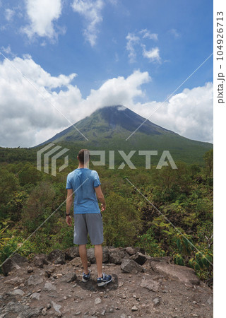 Rear view of male traveler admiring the Arenal volcano and idyllic landscape of Costa Rica mountain Rear view of male traveler admiring the Arenal volcano and idyllic landscape of Costa Rica mountain 104926713