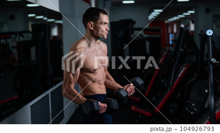 Shirtless man doing bicep exercises with dumbbells in the gym.  104926793